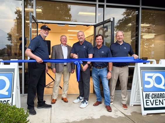 5 men stand behind a large ribbon in front of a building to signify a grand opening.